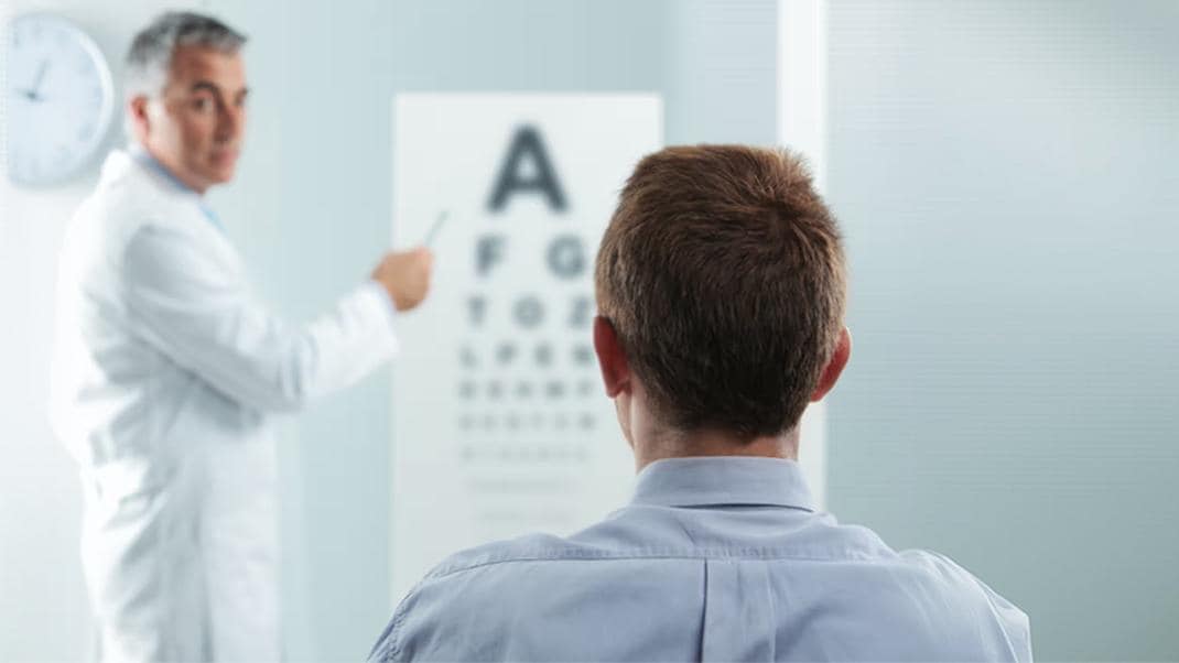 Persona sottoposta a un test della vista davanti a una tabella optometrica, con un professionista sanitario che indica le lettere da leggere durante la visita oculistica.