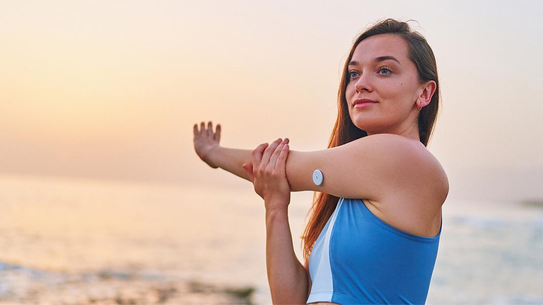 Persona che pratica stretching sulla spiaggia al tramonto indossando un sensore per il monitoraggio continuo del glucosio applicato al braccio, in un contesto che richiama attività fisica e gestione della salute.