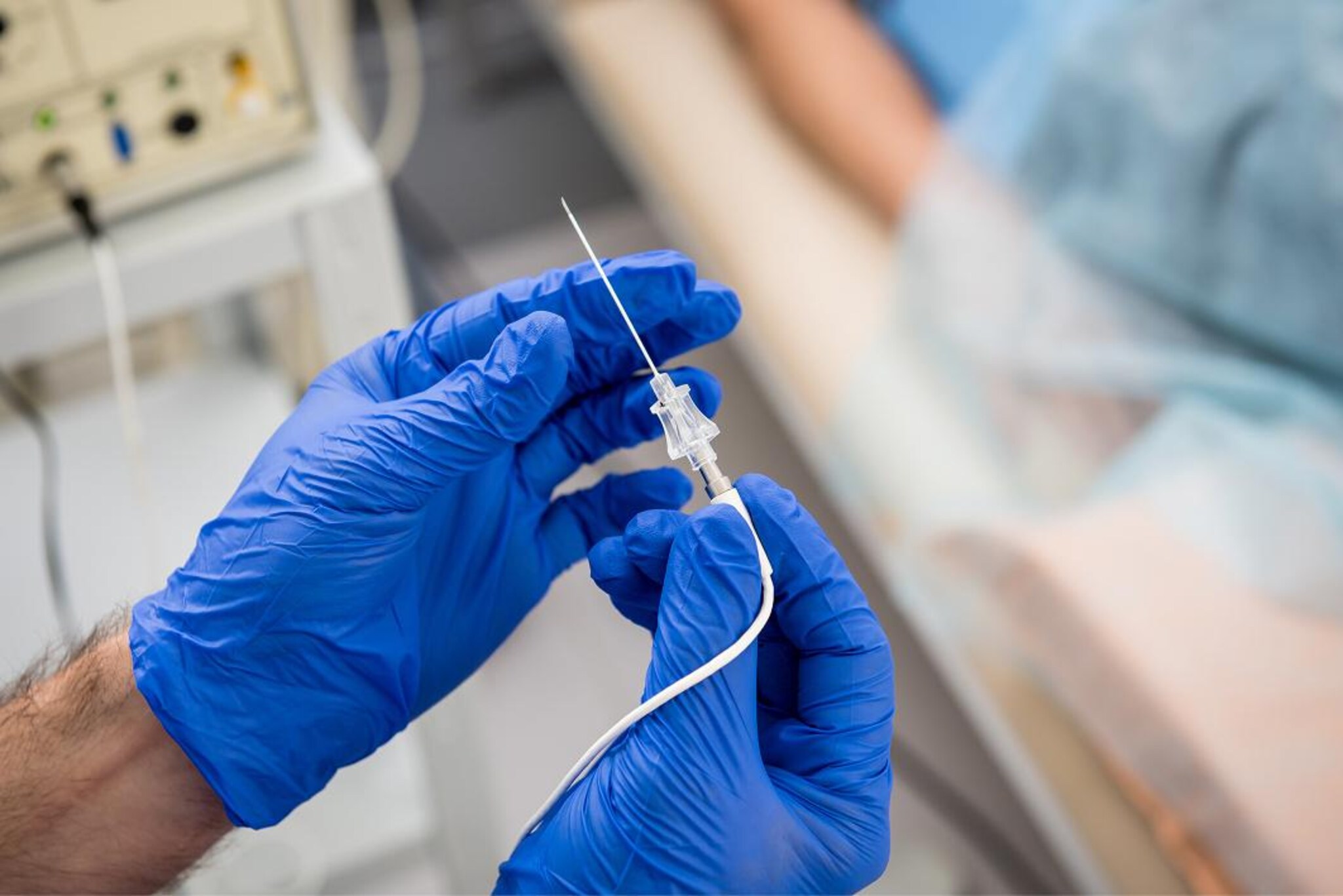 Mani con guanti medicali blu che preparano un sottile ago collegato a un dispositivo medico, con una persona distesa sullo sfondo.