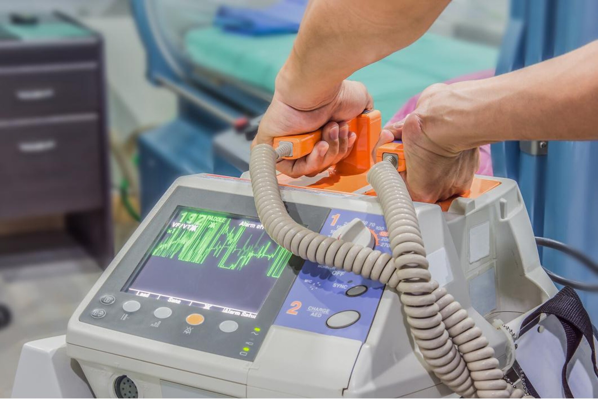 Mani che impugnano le piastre di un defibrillatore, con monitor del dispositivo che mostra un tracciato cardiaco.