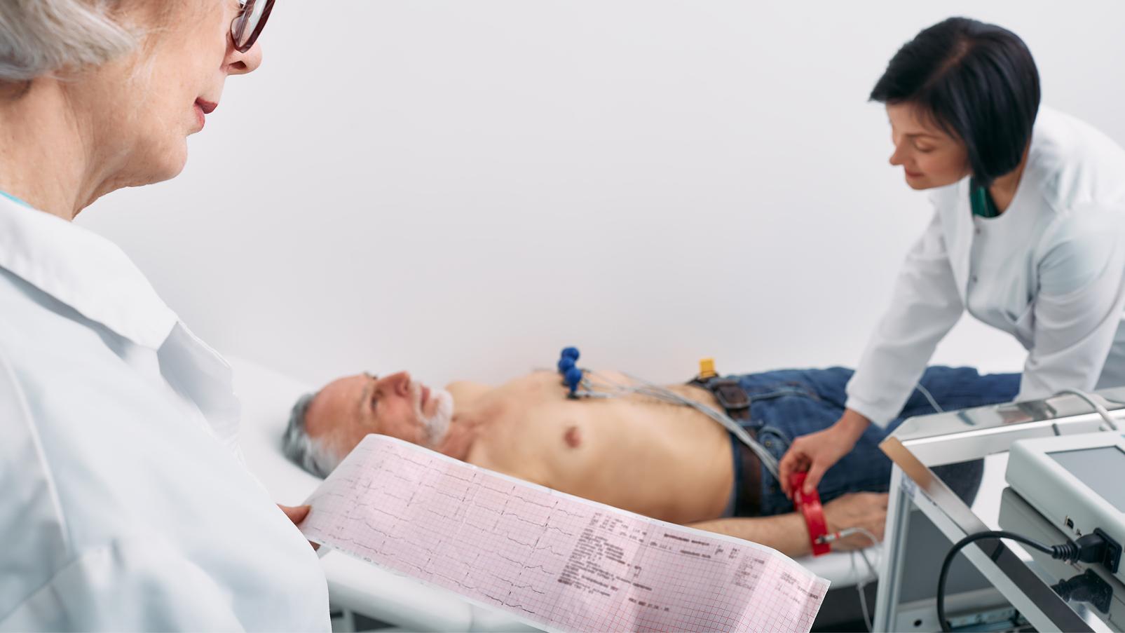 Persona distesa su un lettino durante un elettrocardiogramma, con elettrodi applicati al torace e personale sanitario che controlla l'esame e il tracciato stampato.