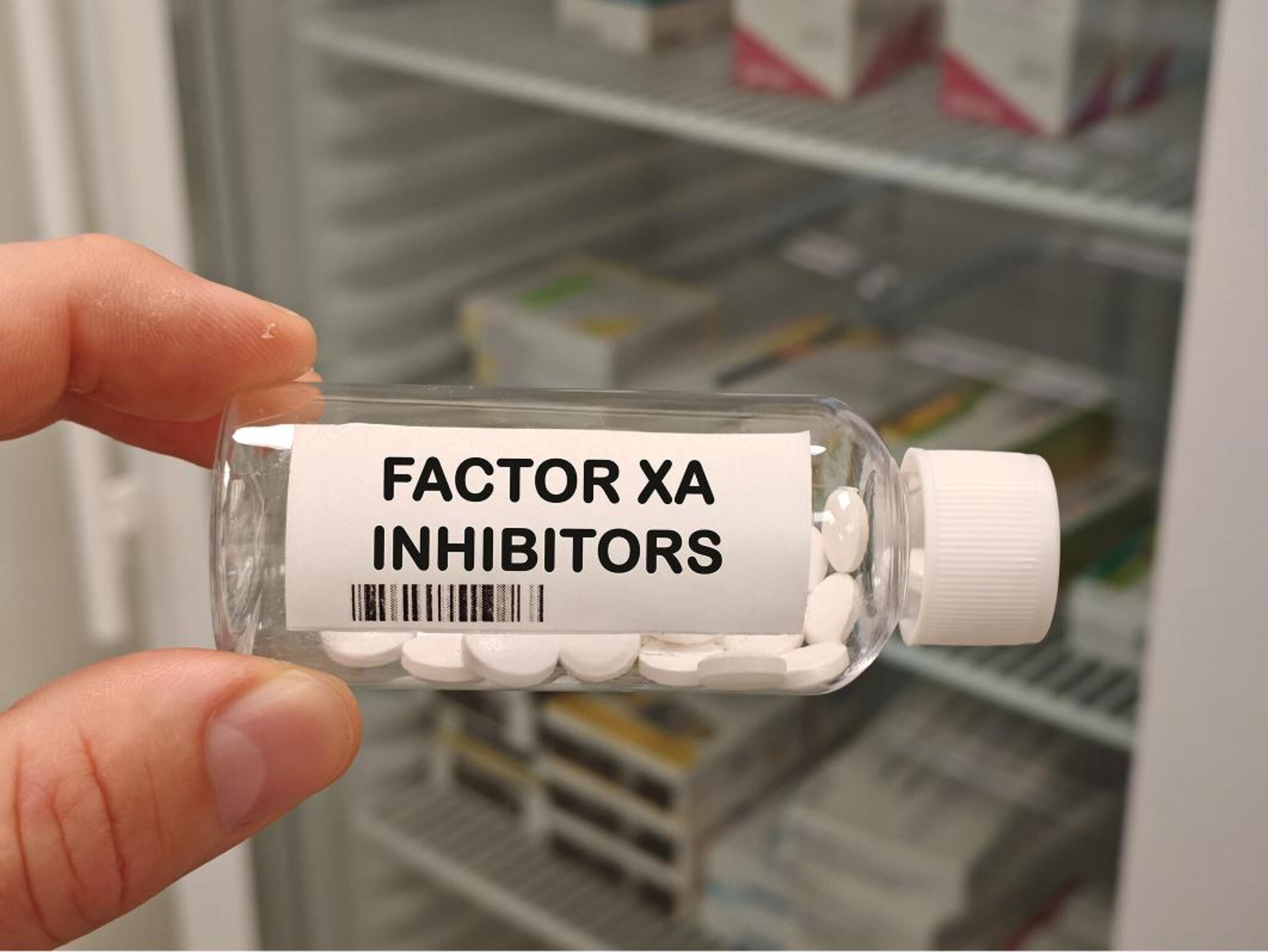 Flacone trasparente contenente compresse bianche, tenuto in mano davanti a un armadietto di medicinali, con etichetta “Factor Xa Inhibitors”.