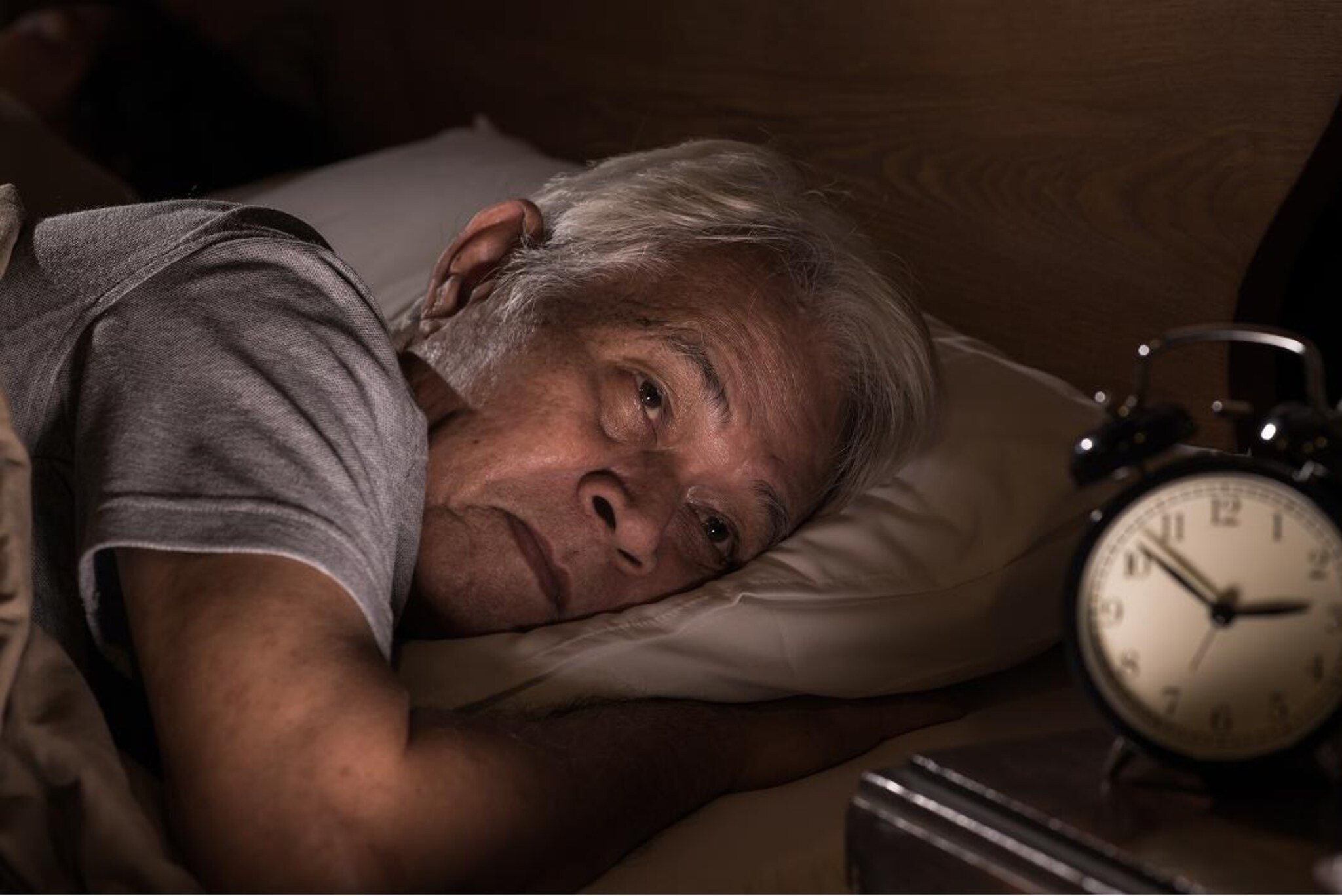 Persona anziana distesa a letto con gli occhi chiusi e la testa appoggiata sul cuscino, in una camera illuminata da una luce calda e soffusa.
