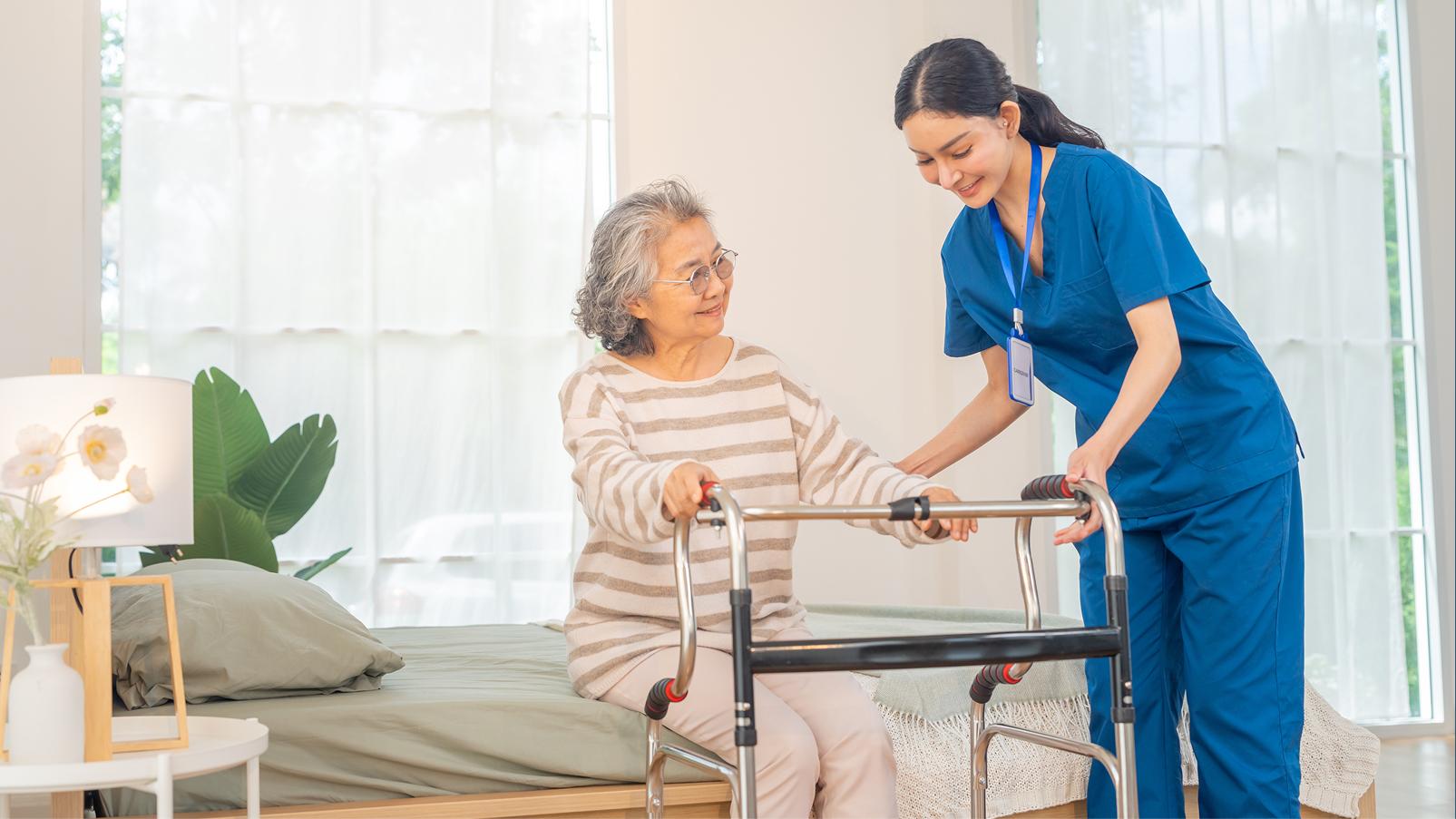 Persona sul bordo di un letto che prova ad alzarsi, sorretta da un deambulatore ed aiutata da un professionista sanitario