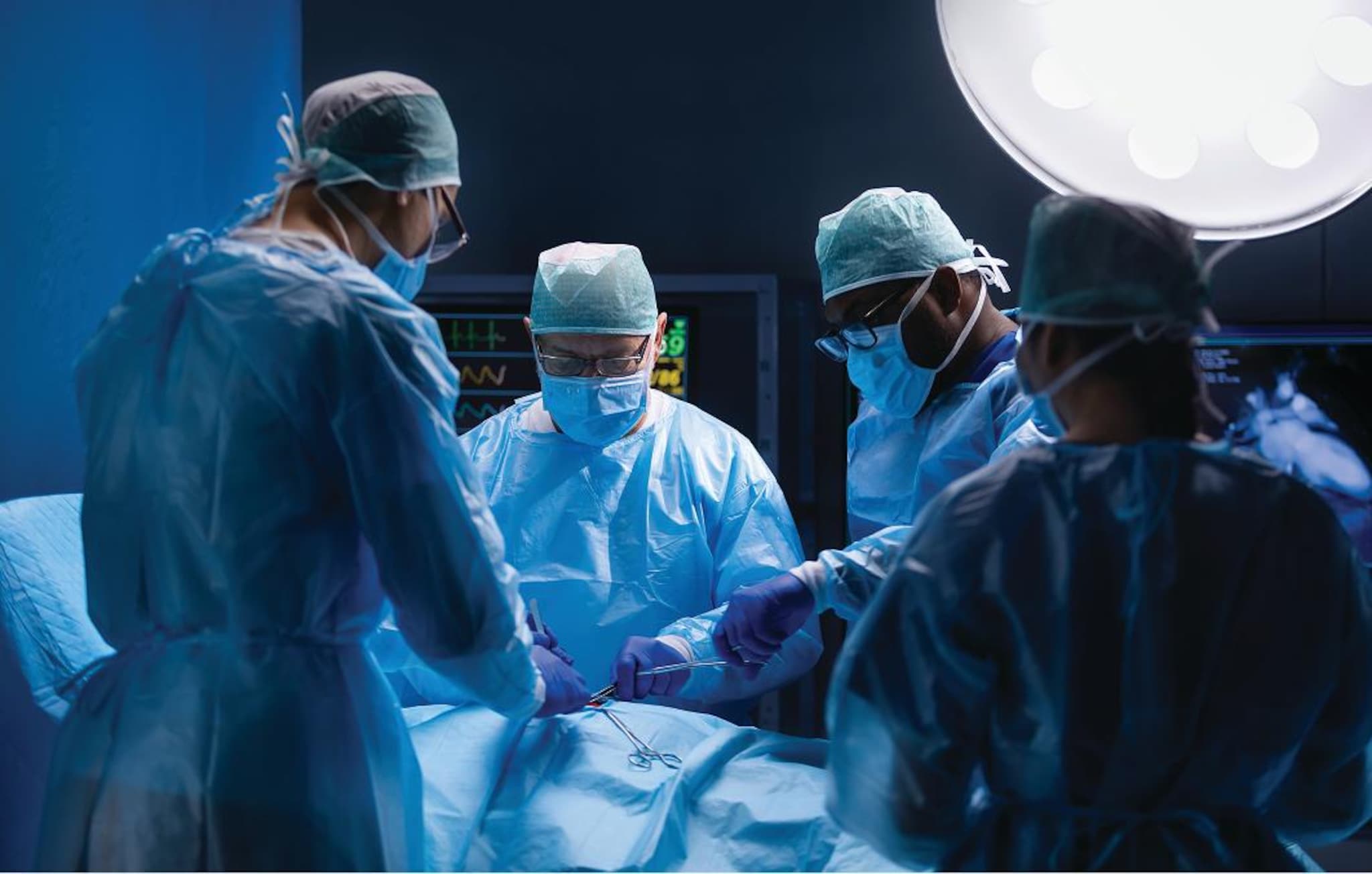 Équipe sanitaria con camici, mascherine, cuffie e guanti esegue un intervento chirurgico in sala operatoria, sotto una lampada chirurgica e con monitor medicali sullo sfondo.