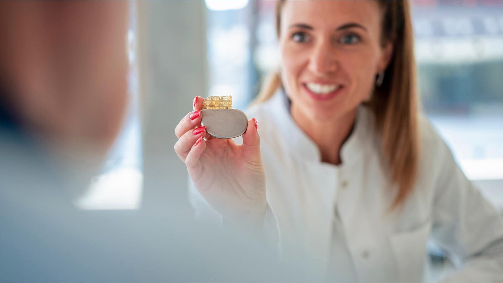 Professionista sanitario in camice bianco mostra a una persona un piccolo dispositivo cardiaco impiantabile tenendolo in mano.