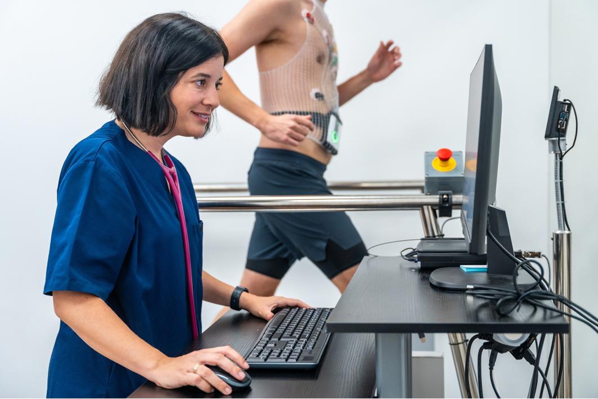 Professionista sanitario controlla un monitor mentre una persona cammina su un tapis roulant con elettrodi applicati al torace e un dispositivo di monitoraggio alla vita.